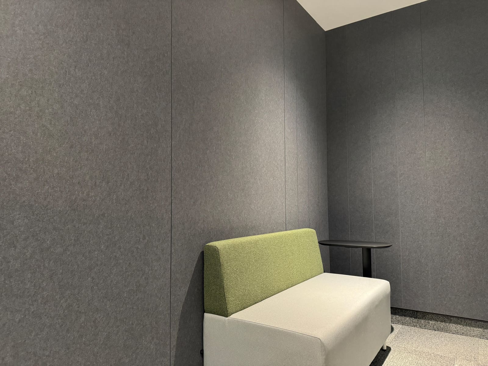 Dark grey ÜNO Board acoustic felt panels covering all walls of a quiet nook with a green-backed white sofa and side table
